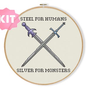 May include: A cross-stitch kit featuring a wooden embroidery hoop containing white fabric with two crossed swords embroidered in gray and purple. The text "STEEL FOR HUMANS" is stitched above the swords and "SILVER FOR MONSTERS" is stitched below.