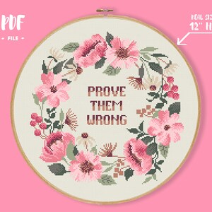 Pode incluir: Um design de ponto cruz num bastidor de 30 cm apresenta uma grinalda floral com flores cor-de-rosa e folhas verdes. O texto "PROVE THEM WRONG" est&aacute; centrado dentro da grinalda. O design est&aacute; sobre um tecido creme, sobre um fundo rosa.
