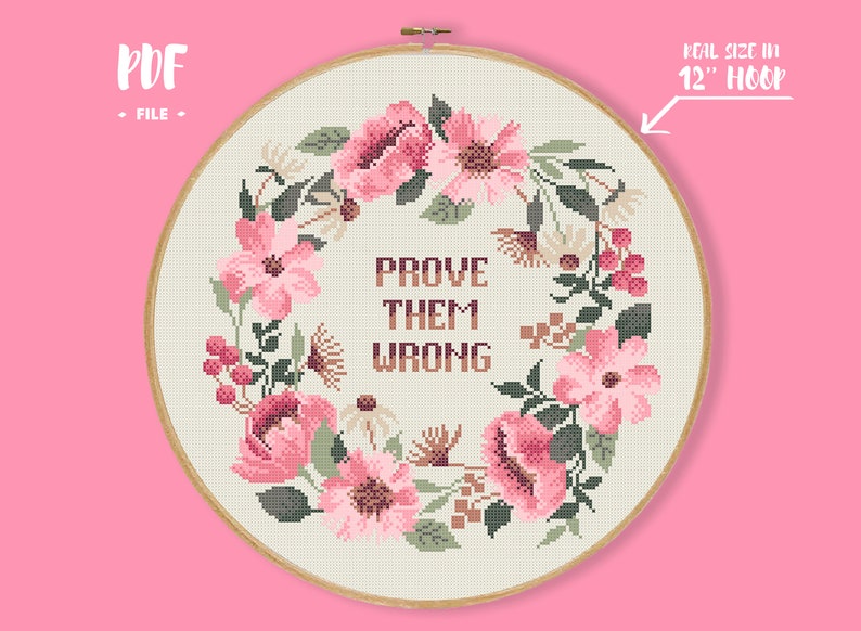 Pode incluir: Um design de ponto cruz num bastidor de 30 cm apresenta uma grinalda floral com flores cor-de-rosa e folhas verdes. O texto "PROVE THEM WRONG" est&aacute; centrado dentro da grinalda. O design est&aacute; sobre um tecido creme, sobre um fundo rosa.