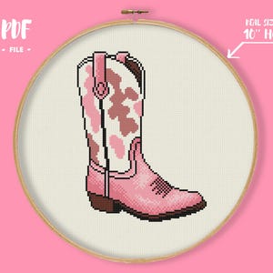 May include: A cross-stitch pattern of a pink cowboy boot with a white and pink cow print design, displayed in a 25.4 cm hoop. The text "PDF FILE" is in the upper left corner. The text "REAL SIZE IN 10" HOOP" is in the upper right corner.
