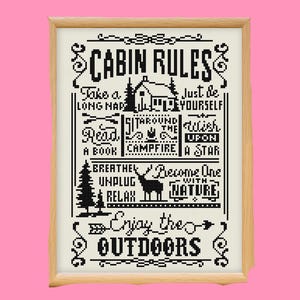 May include: A framed cross-stitch artwork with the words "CABIN RULES" in black on an off-white background. The design includes cabin-themed illustrations and phrases like "Enjoy the Outdoors." The wooden frame adds a rustic touch.