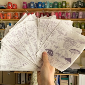 May include: A set of 16 white fabric sheets with various purple line drawings of mushrooms, plants, and a cauldron with a star. The sheets are fanned out in a hand.