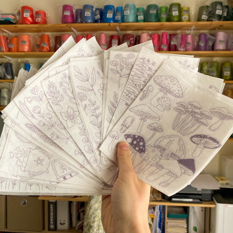 May include: A set of 16 white fabric sheets with various purple line drawings of mushrooms, plants, and a cauldron with a star. The sheets are fanned out in a hand.