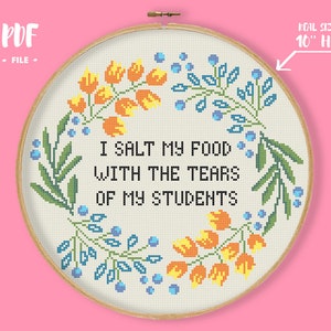 May include: A 10-inch embroidery hoop with a white fabric background featuring a cross-stitch design of a floral wreath with the text "I salt my food with the tears of my students".