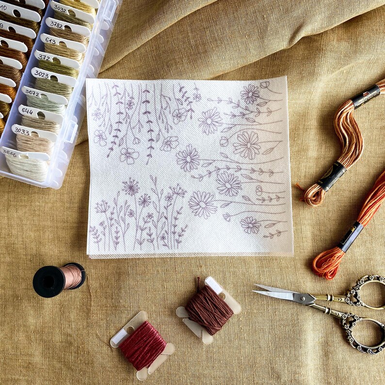 May include: A white embroidery project with a floral design, surrounded by embroidery floss spools in various colors, including brown, orange, and red. The project is on a beige fabric surface.