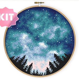 Pode incluir: Um kit de ponto cruz que representa um céu noturno com uma galáxia de estrelas e nuvens sobre uma silhueta de uma floresta. O kit é mostrado em um aro de bordado de madeira.