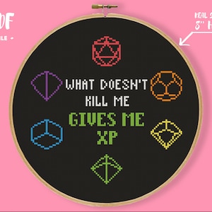 Pode incluir: Um aro de bordado preto com um desenho de ponto cruz que apresenta dados coloridos e o texto "What doesn't kill me gives me XP". O aro tem um diâmetro de 20 cm.