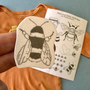 May include: A cream-colored bee-shaped patch with a black and white striped body and wings. The patch is held in front of an orange onesie and a paper with bee illustrations and the text "Stick and Stitch SS28 Bees".