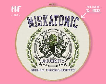 May include: A cross-stitch pattern in a 12-inch wooden hoop featuring the text MISKATONIC UNIVERSITY ARKHAM MASSACHUSETTS and a green tentacled creature emblem surrounded by green laurel leaves on a white fabric background.