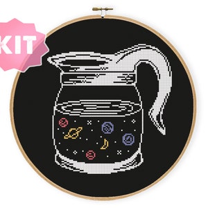 May include: Cross-stitch kit featuring a coffee pot design with a space-themed interior. The pot is white, set against a black background, and framed in a wooden hoop. The interior of the pot includes planets, stars, and a crescent moon.