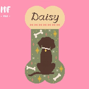 May include: A cross-stitch pattern shaped like a dog bone on a pink background. The top section is cream with the name Daisy in brown text. The green lower section features a brown dog silhouette, white bones, and yellow and pink star accents.