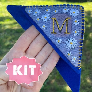 May include: A blue felt pennant with a white daisy embroidery design and the letter "M" in yellow thread. The pennant is labeled "KIT" in a pink circle.