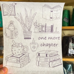 May include: A white fabric coaster with a black and white illustration of books, a plant, a cup of hot chocolate, and a cup of coffee. The text "one more chapter" is printed on the coaster.