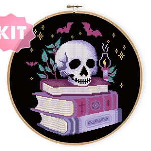Puede incluir: Kit de punto de cruz con una calavera sobre libros, murciélagos y estrellas sobre fondo negro. Incluye una vela, hojas y la palabra "KIT" en una etiqueta rosa. El diseño está en un aro de madera.