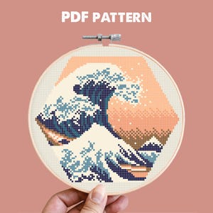 Op de afbeelding: Een borduurring met een pixelontwerp van een golf in blauwe, crème- en perziktinten. De tekst "PDF PATTERN" staat bovenaan. De borduursel wordt in een houten ring gehouden.