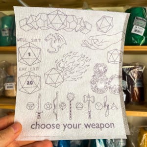Op de afbeelding: Witte keukendoek met een zwart-wit ontwerp met dobbelstenen, een draak, een hand, vuur, een symbool en wapens. De tekst "Well, Shit", "Eat Crit", "Choose Your Weapon" is op de handdoek gedrukt.