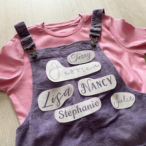 Pode incluir: Uma camisa rosa canelada com gola redonda por baixo de um macacão de veludo roxo. O macacão tem etiquetas com os nomes: Terry, Jessica, Lisa, Nancy, Stephanie e Julie.