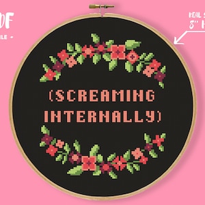 May include: Cross-stitch embroidery in a wooden hoop. The black fabric features the text "(SCREAMING INTERNALLY)" in pink, surrounded by a floral wreath of pink and green cross-stitched flowers. The hoop is 8 inches in diameter.