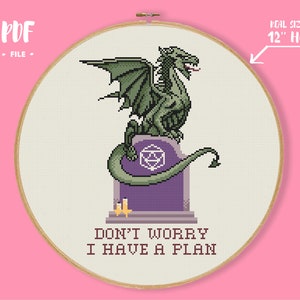 Puede incluir: Patrón de punto de cruz con un dragón verde sentado en una lápida púrpura con el texto "No te preocupes, tengo un plan". El patrón está diseñado para un aro de 30 cm.