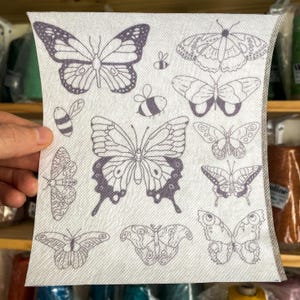 Może przedstawiać: Biały materiał z powtarzającym się wzorem czarnych motyli. Motyle są w różnych rozmiarach i pozycjach, niektóre zwrócone w lewo, a inne w prawo. Na materiale rozrzucone są również małe czarne wzory pszczół.