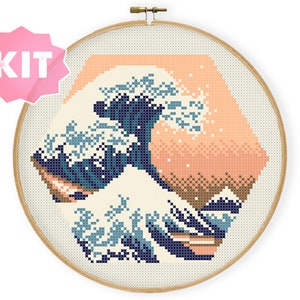Peut inclure: Kit de point de croix représentant une version pixelisée de "La Grande Vague de Kanagawa" de Hokusai. Le motif utilise des nuances de bleu, crème et pêche dans une forme hexagonale, encadrée par un cercle à broder en bois. Le mot "KIT" est dans une étoile rose.