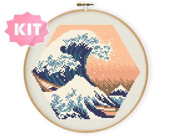May include: Cross-stitch kit featuring a pixelated depiction of Hokusai's "The Great Wave off Kanagawa." The design uses shades of blue, cream, and peach within a hexagonal shape, framed by a wooden embroidery hoop. The word "KIT" is in a pink starburst.