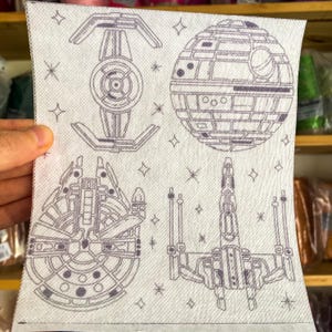 Peut inclure: Un essuie-tout blanc avec quatre illustrations grises de vaisseaux spatiaux et de stations spatiales. Les illustrations comprennent un chasseur TIE, une Étoile de la Mort, le Faucon Millenium et un chasseur X-wing. Le papier essuie-tout est tenu par une main.