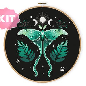 Könnte beinhalten: Kreuzstich-Set mit einem grünen und weißen Motten-Design auf schwarzem Hintergrund. Die Motte ist von Blättern, Monden und Sternen umgeben. Das Design ist in einem Holzreifen gerahmt, mit einem rosa "KIT"-Aufkleber.