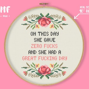 Op de afbeelding: Kruissteekborduurwerk in een houten borduurring. Het ontwerp toont de tekst "ON THIS DAY SHE GAVE ZERO FUCKS AND SHE HAD A GREAT FUCKING DAY" met bloemmotieven. De ring heeft een diameter van ongeveer 25 cm.