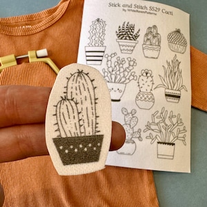 Könnte beinhalten: Ein beiger Kaktusaufnäher mit einer schwarzen Umrandung von zwei Kakteen in einem Topf wird vor einem Blatt mit Kaktusdesigns gehalten. Das Blatt trägt die Aufschrift "Stick and Stitch SS29 Cacti by WhiteRavenPatterns". Ein orangefarbenes Oberteil ist im Hintergrund.