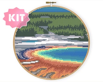 May include: Cross stitch pattern of a colourful landscape with a blue lake, green trees, and a brown and orange rocky shore.