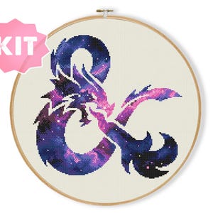 Puede incluir: Kit de punto de cruz con un diseño de dragón y ampersand, que muestra un patrón de galaxia en tonos morados, azules y rosas. El diseño está en un bastidor de bordado de madera. Una pegatina rosa en forma de estrella con "KIT" está en la esquina superior izquierda.