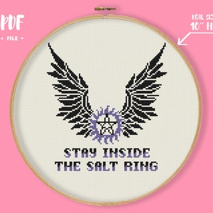 May include: Cross-stitch design in a 10-inch hoop featuring black wings above a purple pentagram and the text "STAY INSIDE THE SALT RING". The design is on a cream-colored fabric, with a wooden hoop and a pink background.