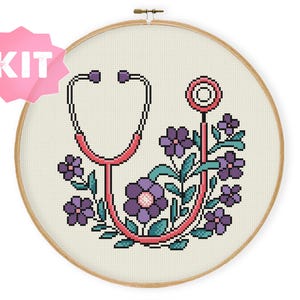 Pode incluir: Um projeto de ponto cruz num bastidor de madeira redondo com um estetoscópio rosa pixelizado entrelaçado com flores roxas e folhas verde-azuladas em tecido branco. Um gráfico rosa em forma de estrela com o texto branco KIT está no canto.