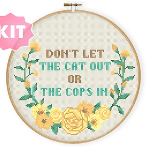 May include: A cross-stitch embroidery kit featuring a floral wreath with yellow flowers and green leaves. The text "DON'T LET THE CAT OUT OR THE COPS IN" is stitched in brown and teal thread.