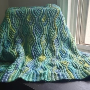 Puede incluir: Una manta de ganchillo con un patrón ondulado y texturizado en tonos verde azulado, turquesa, verde lima y azul claro. El diseño consta de columnas verticales ondulantes con puntos en relieve que crean un efecto dimensional y ondulado.