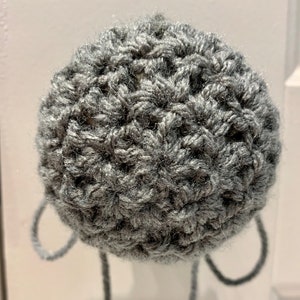 May include: A gray crocheted ball with two loops and a long strand of yarn extending from the bottom. The ball is made of a textured yarn, and the loops and strand are the same color as the ball.