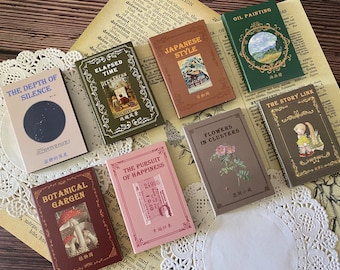 Può includere: Una collezione di dieci piccoli libri decorativi colorati con vari titoli e illustrazioni. I libri sono disposti in cerchio su un pizzo bianco. I titoli includono "The Depth of Silence", "Elapsed Time", "Japanese Style", "The Pursuit of Happiness", "Flowers in Clusters", "Oil Painting", "The Story Link", "Botanical Garden", e altro ancora.