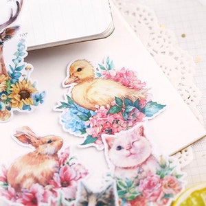 Puede incluir: Un conjunto de pegatinas de acuarela con una variedad de animales y dise&ntilde;os florales. Las pegatinas incluyen un ciervo, un patito, un conejo y un gato, todos rodeados de flores y vegetaci&oacute;n.