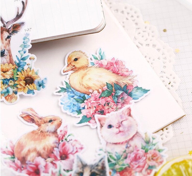 Puede incluir: Un conjunto de pegatinas de acuarela con una variedad de animales y dise&ntilde;os florales. Las pegatinas incluyen un ciervo, un patito, un conejo y un gato, todos rodeados de flores y vegetaci&oacute;n.