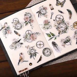 May include: A collection of decorative stickers featuring vintage clocks, gears, flowers, birds, feathers, and a butterfly. The stickers have a soft color palette with touches of gold and are arranged on an open notebook page.