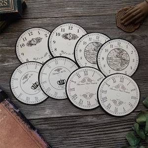 May include: Eight vintage clock face paper craft embellishments with black Roman numeral markings and black borders. The clock faces have a cream background and feature different designs, including a crown, a map, and a floral pattern.