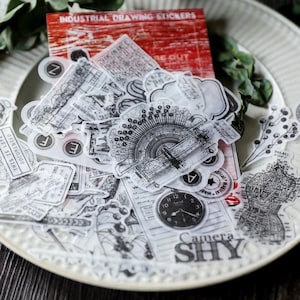 May include: A sheet of clear stickers with black and white illustrations of vintage objects, including a typewriter, a clock, and a camera. The stickers are labeled "Industrial Drawing Stickers".