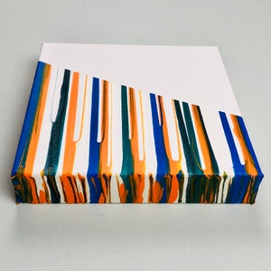 Puede incluir: Pintura abstracta sobre lienzo con rayas verticales de pintura azul, verde, naranja y blanca. Las rayas son irregulares y crean un dise&ntilde;o abstracto din&aacute;mico.