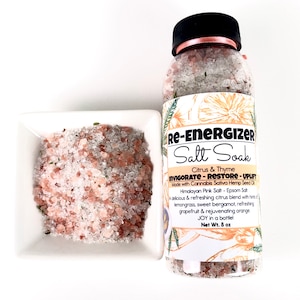 May include: A clear plastic bottle of "Re-Energizer Salt Soak" with a black cap and pink ribbon, next to a small white square bowl filled with pink Himalayan salt. The bottle label reads "Citrus & Thyme" and "Net Wt. 8 oz."