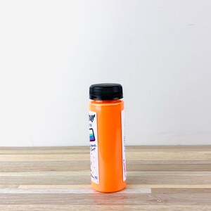 Puede incluir: Una peque&ntilde;a botella de pintura naranja con tapa negra. La botella est&aacute; etiquetada con "Colour" y tiene una franja de arco&iacute;ris en el lateral.