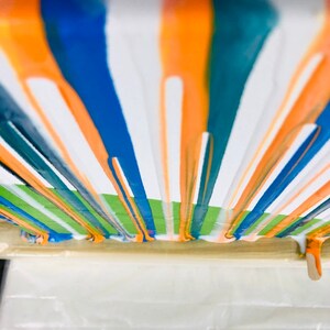 Puede incluir: Una vista cercana de una pintura con rayas verticales de pintura naranja, azul, verde azulado y blanca. La pintura gotea por el lienzo, creando un dise&ntilde;o din&aacute;mico y abstracto.