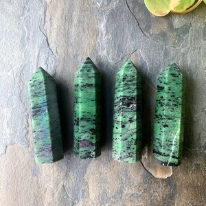 Könnte beinhalten: Vier grüne und schwarze Zoisit-Kristalltürme mit poliertem Finish. Die Kristalle sind in einer Reihe angeordnet und zeigen ihre natürlichen Muster und Farben. Die Kristalle liegen auf einer grauen Steinoberfläche.