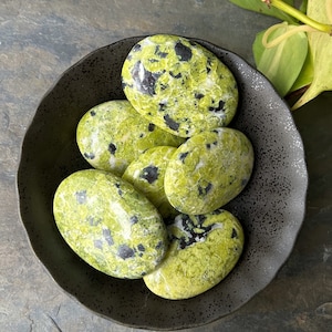 May include: A collection of polished, oval-shaped stones in a black bowl. The stones are a vibrant green color with black speckles. The bowl is dark gray and has a textured surface. The background is a dark gray surface with green leaves.