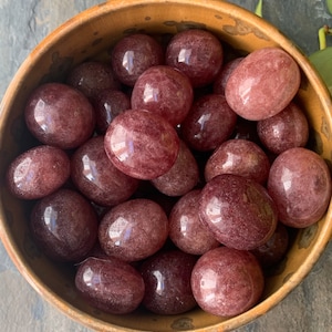 May include: A wooden bowl filled with polished, oval-shaped strawberry quartz stones. The stones are a translucent, reddish-pink color with subtle internal patterns. The bowl has a warm, earthy tone, contrasting with the stones' vibrant hue. The arrangement suggests a collection of gemstones.